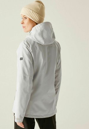 Kobieta w beżowej dzianinowej czapce, ubrana w jasnoszarą kurtkę przeciwdeszczową z kapturem i czarne spodnie, stojąca bokiem na jednolitym tle.