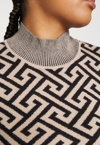 Pull beige avec un col côtelé haut présentant un motif géométrique noir contrasté, texture douce et silhouette ajustée.