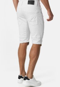 Shorts en denim blanc avec une coupe à hauteur du genou, dotés de deux poches arrière et d'une étiquette en cuir à la taille, assortis à des baskets blanches.