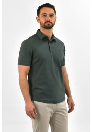 Man staat, draagt een donkergroen polo met korte mouwen en lichtbeige broek, iets naar links gericht tegen een witte achtergrond.