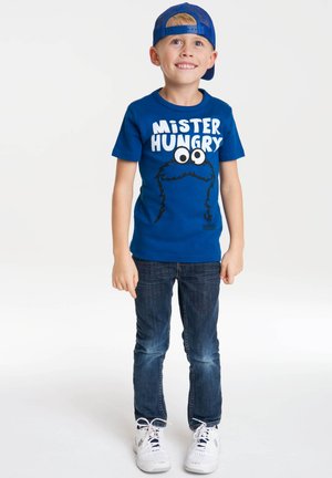 Blue short-sleeve t-shirt with "MISTER HUNGRY" text and cartoon character print. Paired with dark blue jeans and white sneakers.