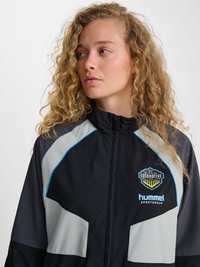 Blondine met krullend haar die een zwart-grijze jas met rits draagt met de logo’s van El Paso Locomotive FC en Hummel Sportswear, kijkt naar links.
