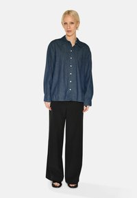 Camicia in denim scuro con colletto e taschino sul petto, abbinata a pantaloni neri larghi e sandali piatti.