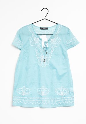 Lichtblauw blouse met korte mouwen en witte geborduurde bloemmotieven, vooraan met kralen gestrikt, hangend aan een zwarte kledinghanger.
