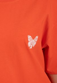 Oranžové bavlněné tričko s kulatým výstřihem, s bílou grafikou motýla a textem na levé části hrudi. Měkká, hladká textura.