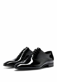 Chaussures habillées en cuir verni noir avec une finition lisse, des pointes effilées et un design de laçage complexe. Talon bas, silhouette élégante.