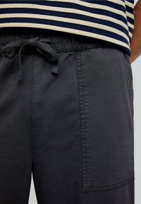 Pantaloni elastici de culoare gri închis, ușori, cu talie elastică și șnur, având o buzunar lateral și o textură netedă. Top cu dungi vizibil deasupra.