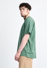 Groene katoenen T-shirt met korte mouwen, ronde hals en zijnadeling. Model draagt beige cargobroek. Achtergrond is eenvoudig wit.