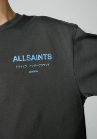 Tamsiai pilkas medvilninis marškinėliai su mėlynu siuvinėtiniu tekstu: "ALLSAINTS," japoniškais simboliais ir "LONDON" šalia apačios. Trumpomis rankovėmis.