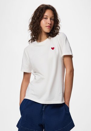 Mlajša ženska s kodrastimi lasmi, oblečena v belo majico s majhnim rdečim srcem na prsih in temno modre kratke hlače, roke v žepih.