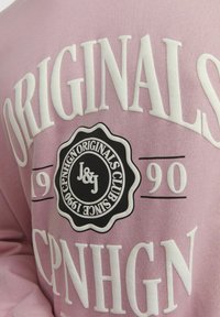 Sudadera de color rosa claro con texto en negrita blanco. Logo circular negro central con detalles de "J&J" y "CPNHGN ORIGINALS CLUB SINCE 1990". Tejido suave.
