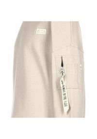 Beżowa bluza z suwakiem umieszczonym na rękawie, materiałowa metka z napisem "USUŃ PRZED LOTEM" oraz naszywka z logo na ramieniu.