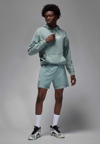 Lys grøn bomulds-hoodie med en kengurulomme, parret med matchende shorts. Modellen har hvide strømper og sort-hvide sportssko på.