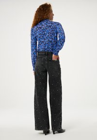 Schwarze Jeans im Weitleg-Design mit Perlenakzenten, kombiniert mit einer blauen Bluse aus transparenter, floral gemusterter Stoffqualität. Der Stoff wirkt leicht und strukturiert.