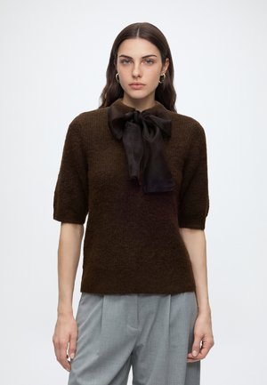 Kvinde med langt mørkt hår iført guldfarvede hoop-øreringe, en mørkebrun kortærmet sweater med en stor sort sløjfe og lysegrå bukser.