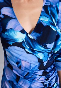 Robe florale bleue avec un décolleté en V et une texture froncée. Fabriquée dans un tissu lisse avec de grands motifs de fleurs bleues claires et foncées.