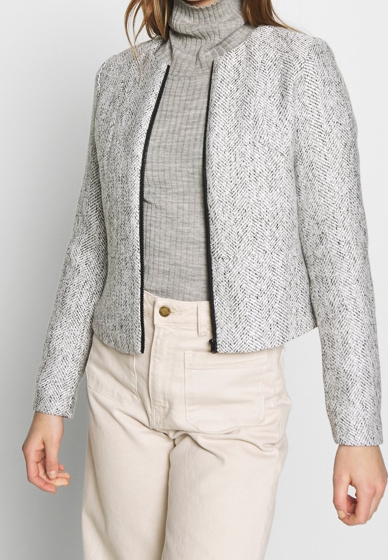 Veste grise à motif chevrons avec une coupe courte, fermeture éclair noire, et manches longues, portée sur un col roulé gris côtelé.