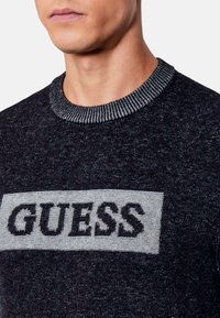 Suéter de punto negro con un logotipo rectangular gris que lleva la palabra "GUESS" en letras negritas, contrastado por un cuello acanalado.