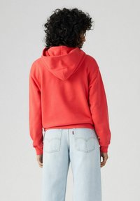 Felpa rossa con cappuccio a coulisse e tasca a canguro frontale, abbinata a jeans azzurri a gamba dritta con tasche posteriori.