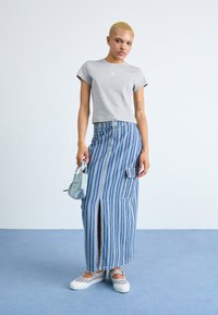 T-shirt cinzento cropped, saia longa azul e branca às riscas com uma fenda, e sapatos brancos com riscas horizontais. Pequena mala azul.