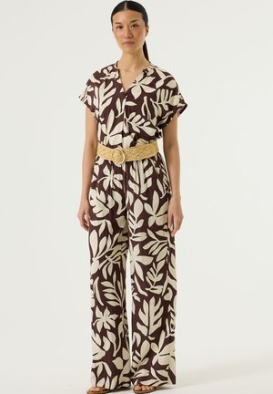 Vrouw staand in een bruine jumpsuit met grote crèmekleurige bladdessin, korte mouwen, wijde pijpen en een brede gevlochten lichtbruine riem tegen een effen achtergrond.