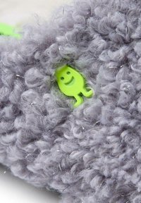 Tessuto grigio ricci con una texture pelosa presenta un'accento a forma di faccina sorridente in plastica verde. Il design include piccoli dettagli riflettenti in tutto il tessuto.