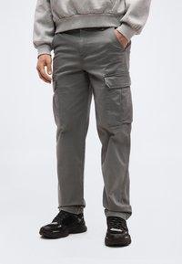 Άνδρας που φορούσε γκρι παντελόνι cargo με τσέπες με καπάκια και μαύρα αθλητικά παπούτσια, στέκεται με το ένα χέρι στην τσέπη μπροστά σε απλό φόντο.