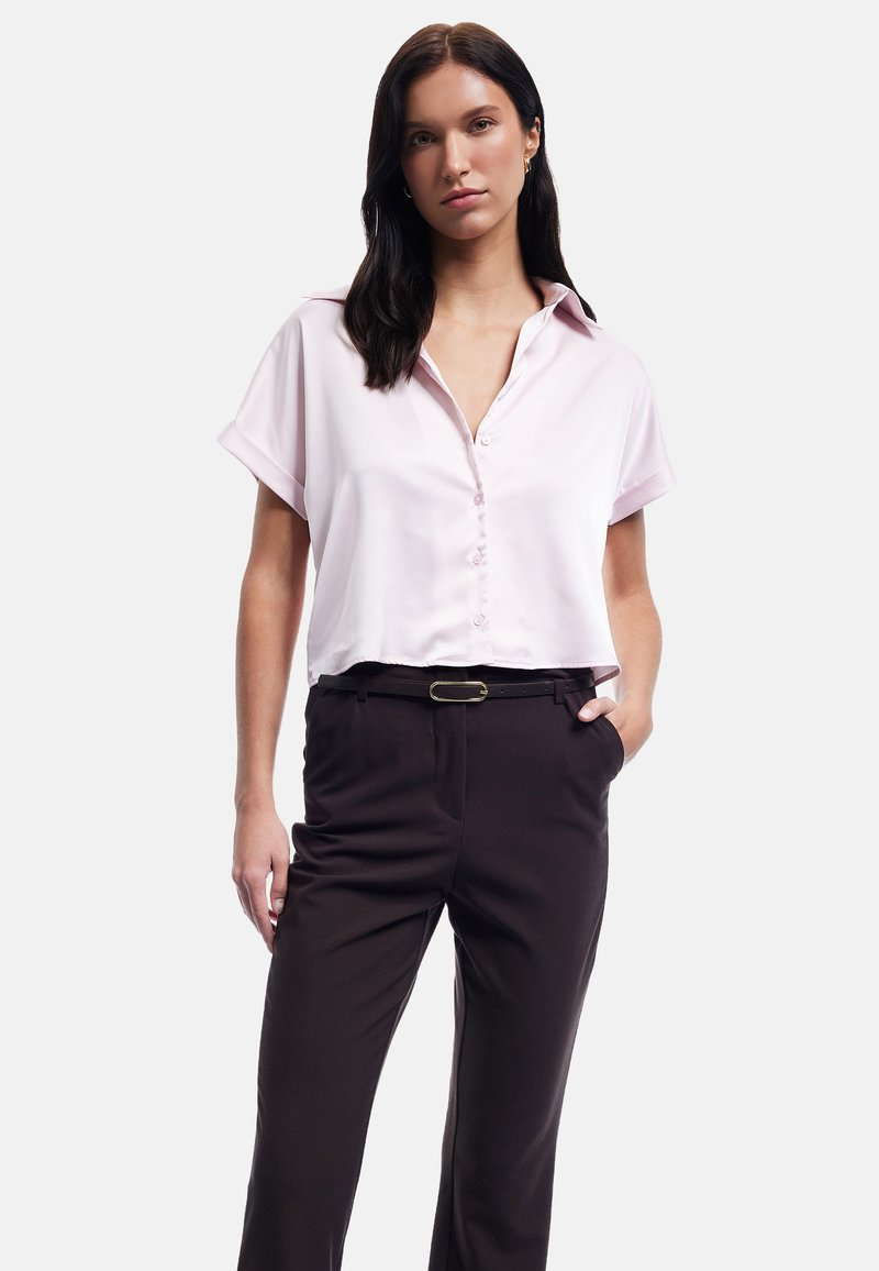 Blusa corta a maniche corte di colore rosa chiaro con colletto e bottoni frontali, abbinata a pantaloni scuri a vita alta di colore marrone scuro e una cintura nera.