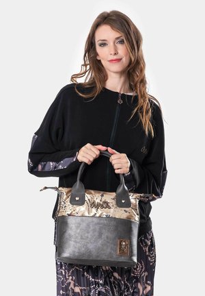 Mujer con cabello ondulado y largo sosteniendo un bolso Anekke negro y beige, vistiendo una sudadera negra con cierre y una falda estampada, frente a un fondo blanco.