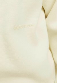 Tissu couleur crème avec une surface texturée. Présente un logo embossé subtil "Originals Studio" en beige clair.