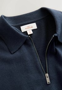 Námořnický modrý úpletový polo s řetězovým zapínáním a měkkým límcem, který má stříbrný zip a bílou etiketu s nápisem "NEXT SIGNATURE" a velikostí "M". Hladká textura.