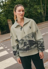 Pulover din fleece de culoare verde deschis, cu fermoar pe un sfert, prezentând un imprimeu cu peisaje negre și păsări. Mansetele și tivul ribb sunt accentuate pentru un plus de textură.