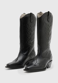 Svarta läderboots i cowboystil med spetsig tå, präglade flammmönster och låg staplad klack. Har en klassisk västerländsk silhuett.