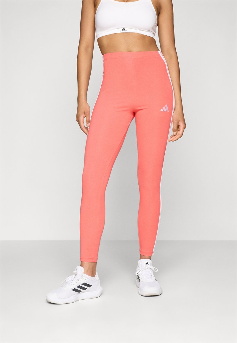 Κοραλλί αθλητικά κολάν με ψηλή μέση, λευκές πλευρικές λεπτομέρειες και λογότυπο Adidas στο αριστερό μέρος του μηρού, συνδυασμένα με λευκά αθλητικά παπούτσια.