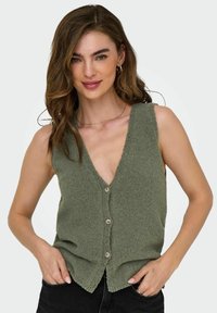 Gilet senza maniche verde oliva con scollo a V, tessuto texturizzato, chiusura frontale con bottoni e orlo sfilacciato. Indossato sopra jeans neri.