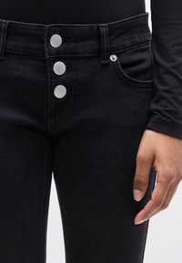 Μαύρο τζιν με ψηλή μέση που διαθέτει τρία μεταλλικά κουμπιά, μπροστινές τσέπες και υφή υφάσματος ντένιμ. Κοντινό πλάνο μέσης και χεριού.