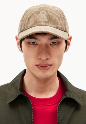 Beige corduroy cap with a slightly curved brim, featuring an embroidered logo on the front panel, worn with a dark green jacket and red shirt.