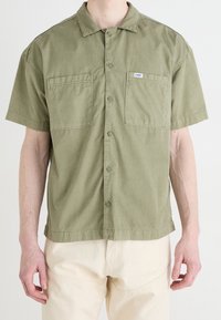 Camicia a maniche corte verde oliva in cotone, con design a bottoni, due tasche sul petto e una piccola etichetta sulla tasca sinistra.