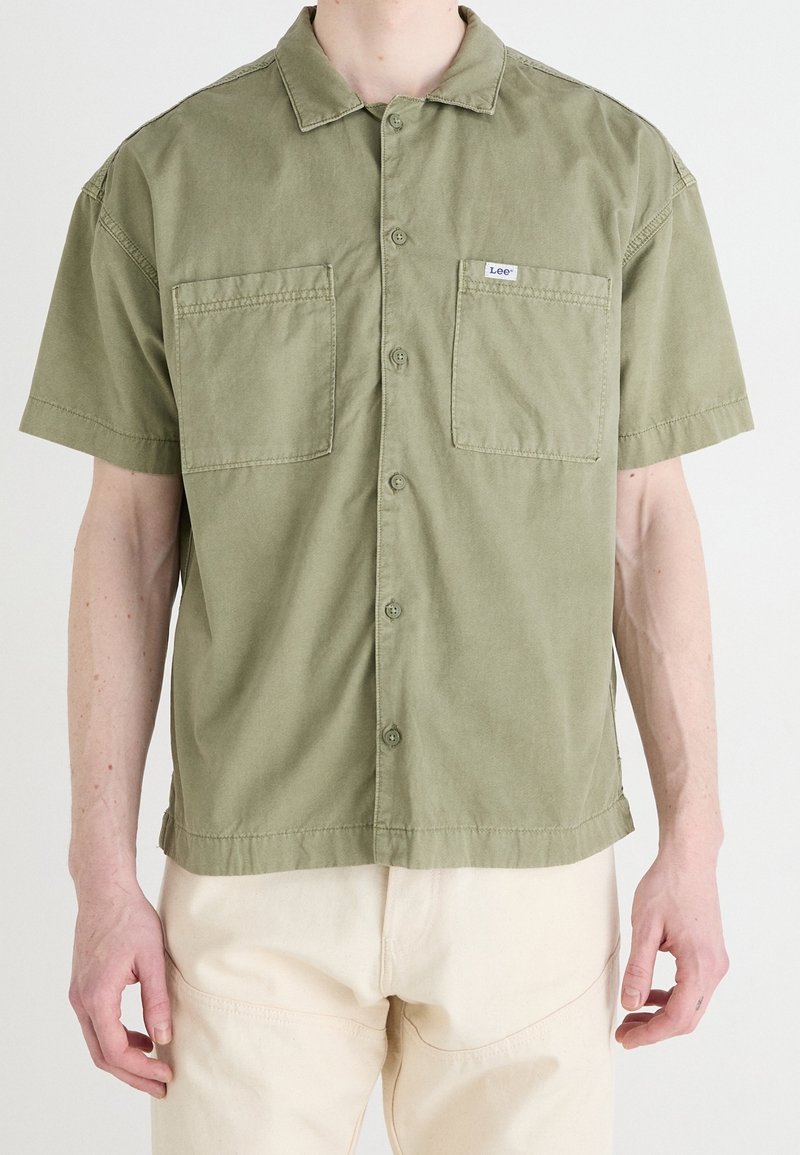 Camicia a maniche corte verde oliva in cotone, con design a bottoni, due tasche sul petto e una piccola etichetta sulla tasca sinistra.