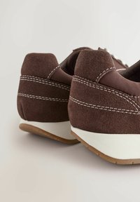 Des baskets en daim marron avec des surpiqûres blanches et une semelle en caoutchouc présentent un design élégant et un talon arrondi. Ornées d'une base blanche contrastante.
