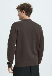 Brauner gestrickter Pullover mit langen Ärmeln, einem gerippten Saum und einem schlichten Kragen, der einen strukturierten Stoff und ein seitliches Logo-Tag aufweist.