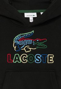 Čierna mikina s kapucňou s farebným vyšitým motívom krokodíla a slovom "LACOSTE" v viacfarebných písmenách. Materiál pôsobí mäkko a štruktúrovane.