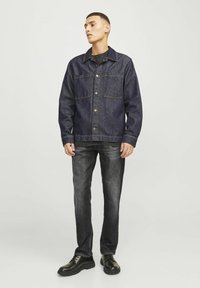 Jeans jakna temno modre barve z dvema sprednjima žepoma, kovinskimi gumbi in kontrastnim šivom; v kombinaciji s ozkimi črnimi kavbojkami in črnimi čevlji.
