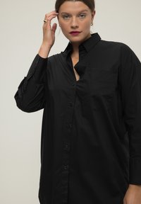 Chemise noire à manches longues en tissu lisse, dotée d'une poche poitrine, de poignets à boutons et d'une coupe droite.