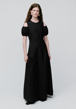 Mujer con cabello rizado, vestida con un largo vestido negro con mangas abullonadas descubiertas en los hombros, de pie contra un fondo claro y liso.