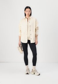 Veste en velours côtelé beige clair avec des boutons, deux poches avant, portée sur un top beige et un legging noir ; tenant un petit sac beige.