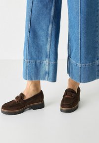Brown suede loafers with a strap and buckle detail, paired with wide blue denim trousers. The shoes feature a textured sole.