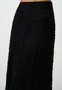 Jupe en dentelle noire avec un motif complexe, texture légère et silhouette fluide, présentant une taille haute et une finition translucide.