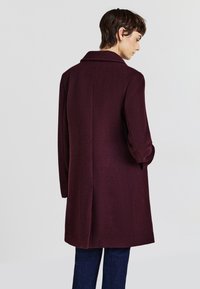 Burgundy wool coat with a classic collar, long sleeves, back vent, and a tailored fit, paired with dark denim trousers.