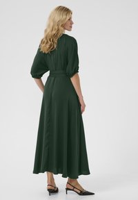 Lungo abito verde scuro con maniche a sbuffo e vita aderente, realizzato in tessuto morbido e fluido, con una gonna svasata. Scarpe nere con tacco a rocchetto visibili.